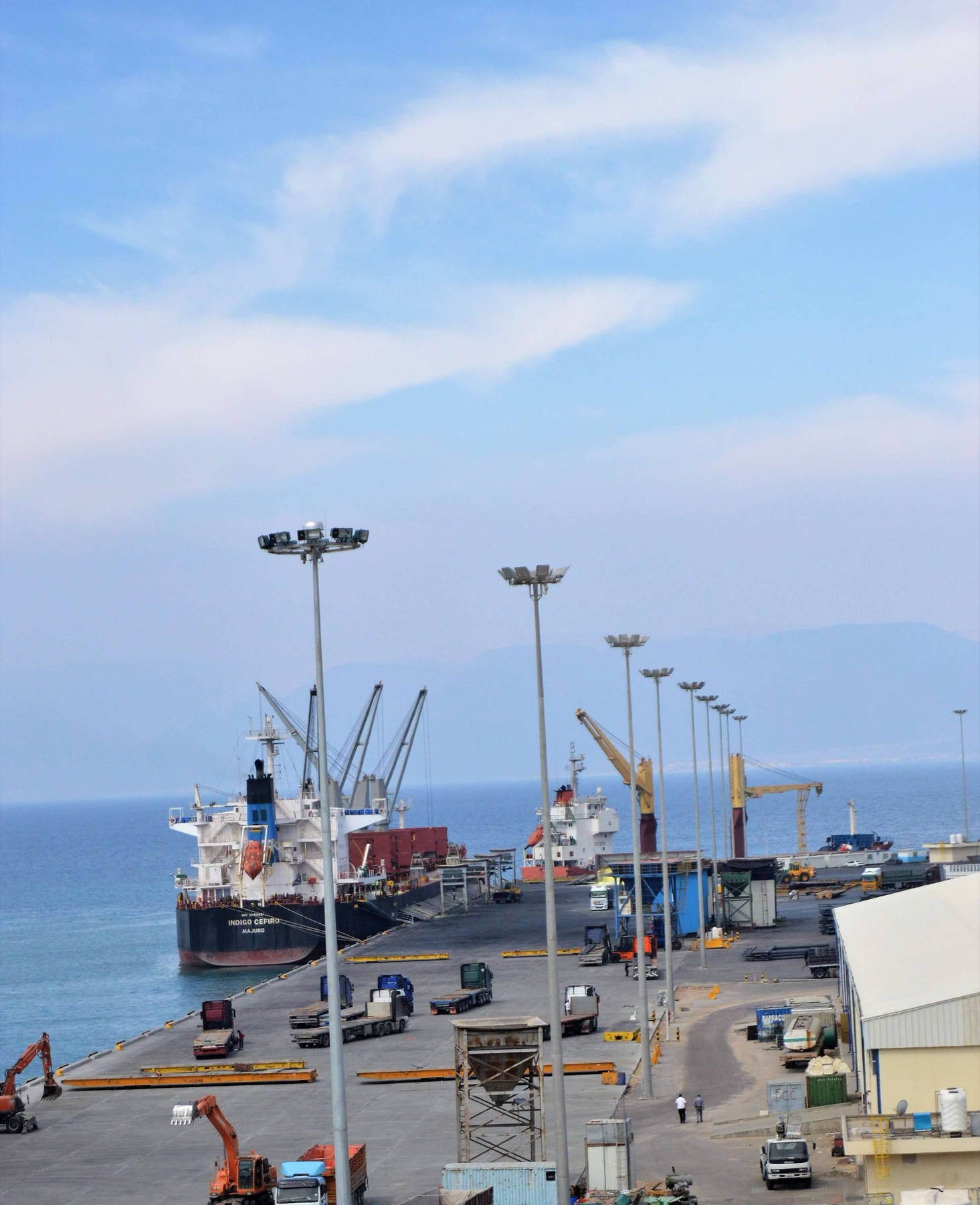 Aqaba port operations with two bulk and general-cargo vessels alongside working quays, mobile cranes, trucks, and the Gulf of Aqaba beyond