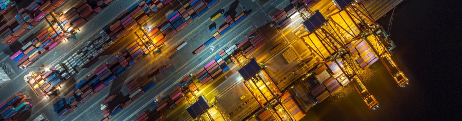 Aerial night view of an ICTSI container terminal with quay cranes, illuminated berths, and container stacks