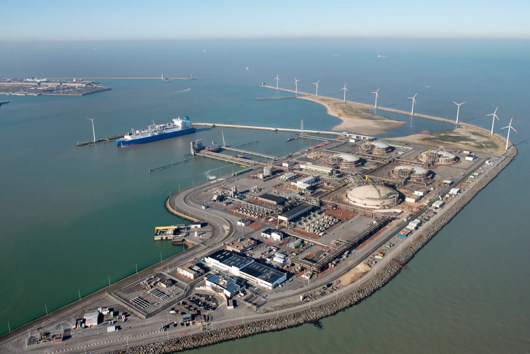 Aerial view of the Zeebrugge port area with an LNG carrier, marine loading jetties, storage tanks, breakwaters, channel entrance, and coastal wind turbines