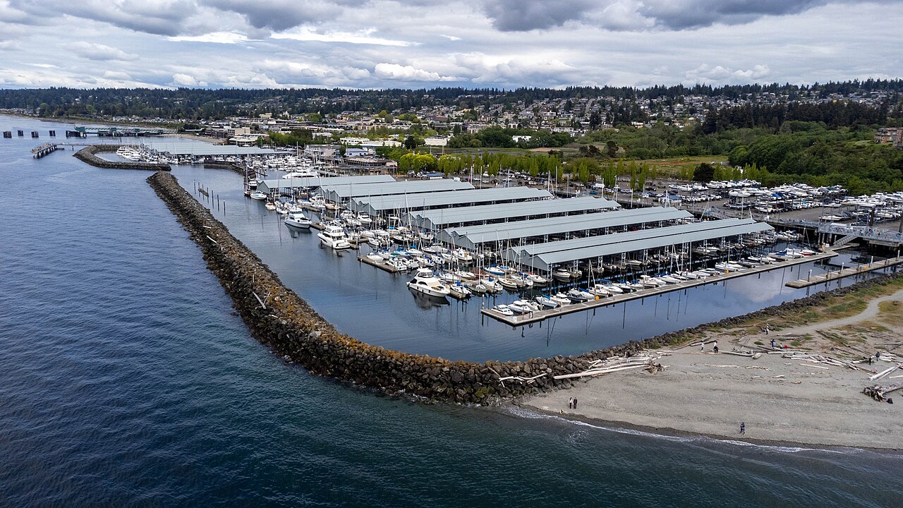 Aerial view across Puget Sound toward the Port of Edmonds marina, showing its rock breakwater, covered and open moorage, dry storage and the Edmonds shoreline
