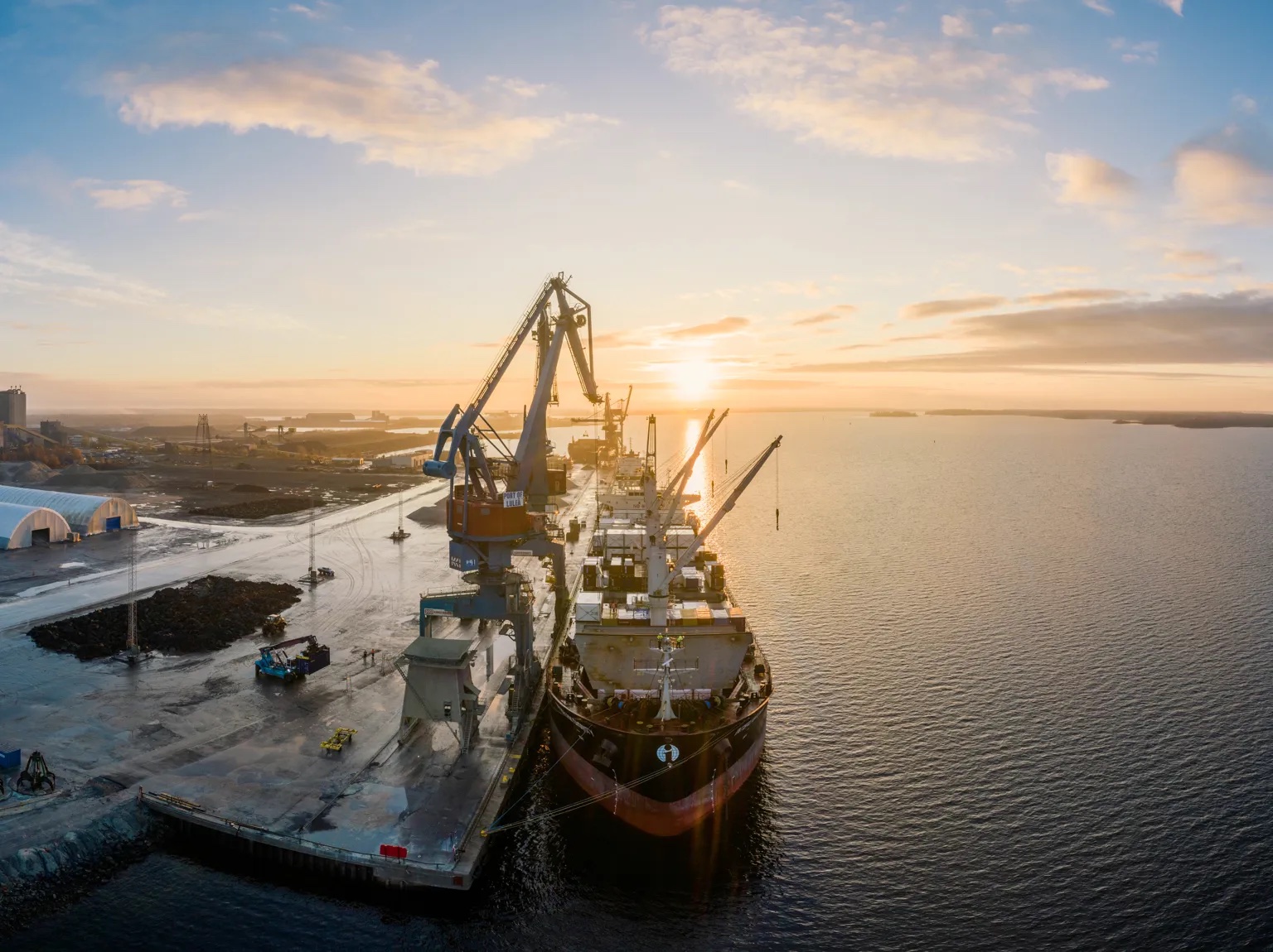 Aerial view of a bulk carrier alongside an operating Port of Luleå quay with ship loaders, mobile equipment, stockpiles, and the Gulf of Bothnia beyond