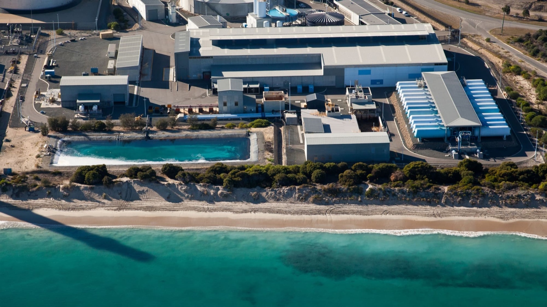 Aerial view of the Perth seawater desalination plant beside the coast, with process buildings, pipe galleries, and clear-water basins